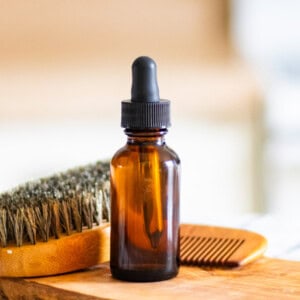 A bottle of homemade beard oil with a beard brush on a wooden board.