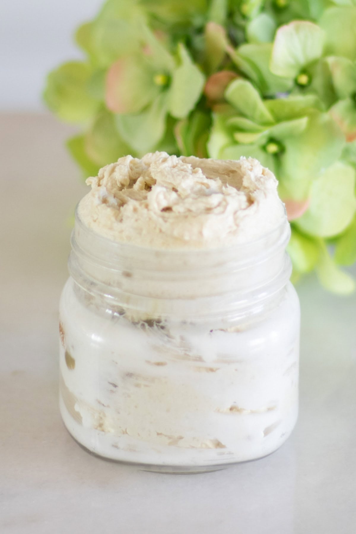A glass jar of homemade body butter with green flowers in the background.