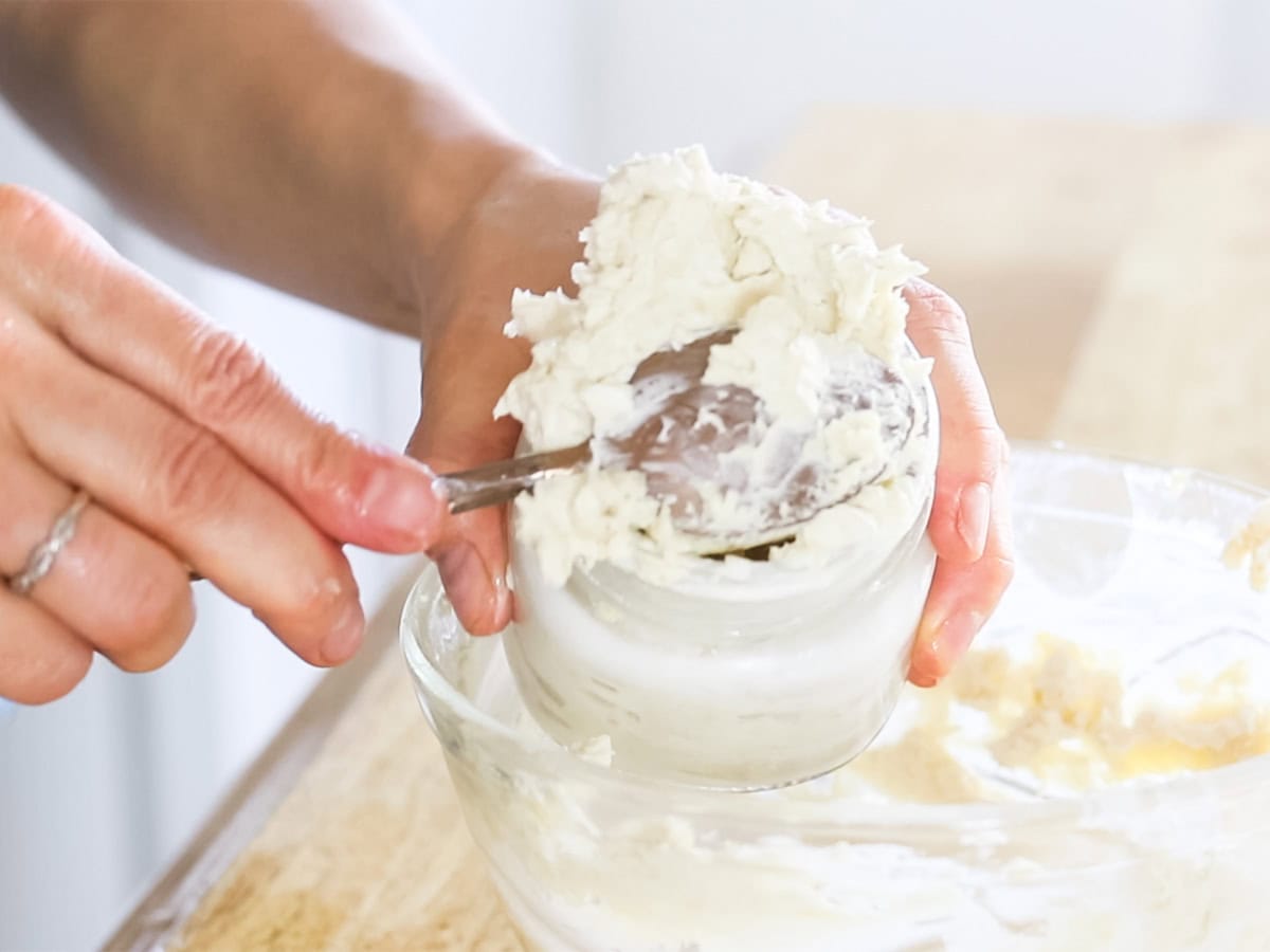 Scooping homemade body butter into a glass jar with a spoon. 