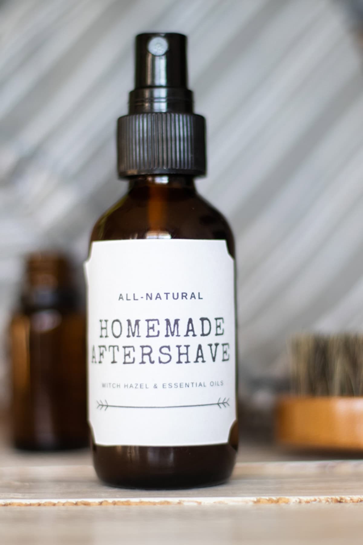 A bottle of homemade aftershave with a printed label next to a brush and bottles of essential oils.