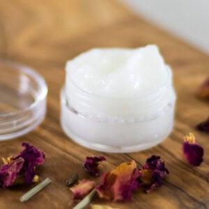 A small jar of eye cream on a wooden table with rose petals.