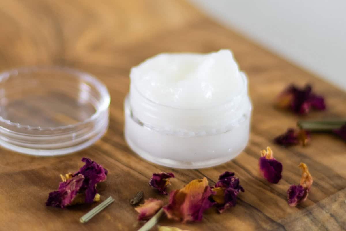 A small jar of eye cream on a wooden table with rose petals.