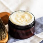 A jar of beard conditioning balm on a striped towel with a brush.