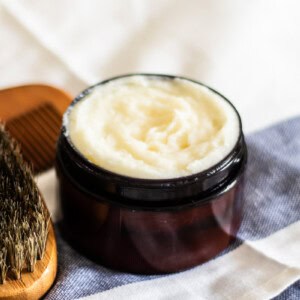 A jar of beard conditioning balm on a striped towel with a brush.