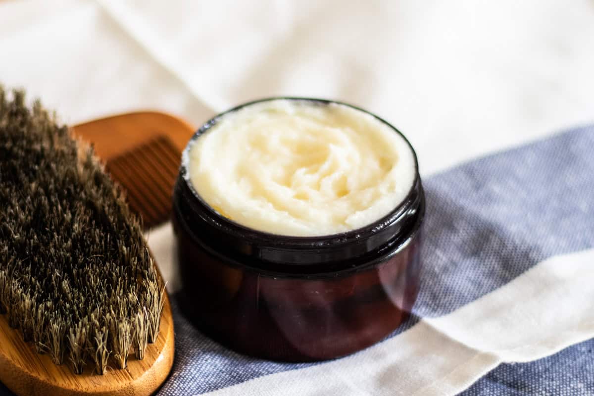 A jar of beard conditioning balm on a striped towel with a brush. 