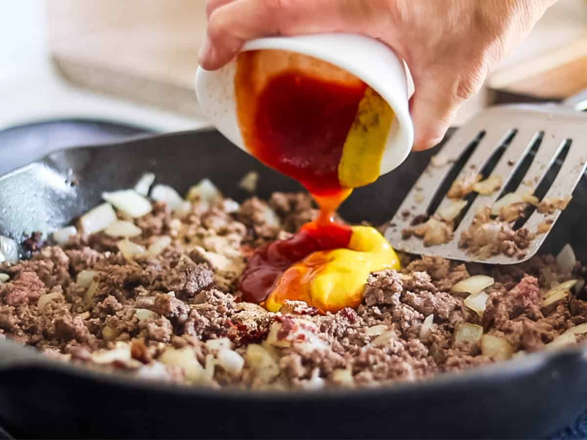 Adding ketchup and mustard to a skillet of ground beef. 