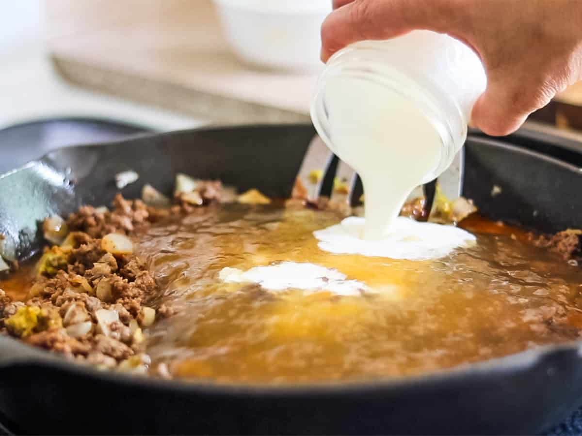 Pouring cream into a skillet with beef and broth to make homemade gluten-free hamburger helper. 
