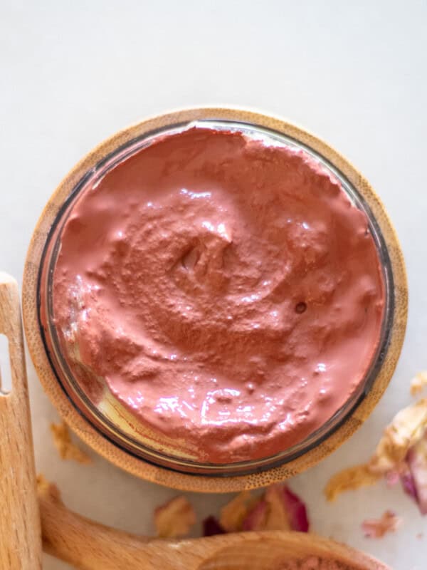 A bowl of rose clay face mask with rose petals and a wooden spoon.