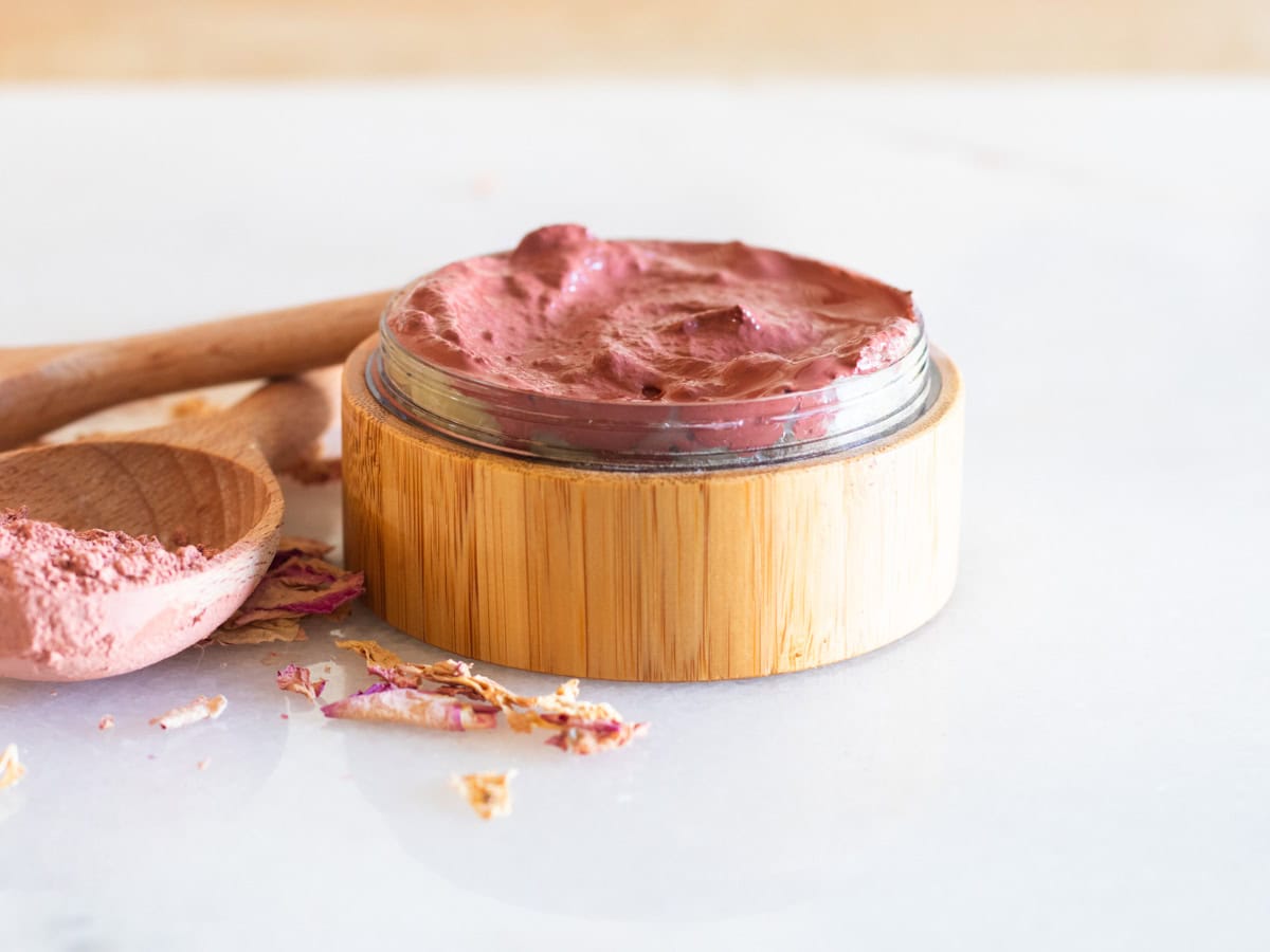 A wooden container of rose clay face mask with a wooden spoon and rose petals. 