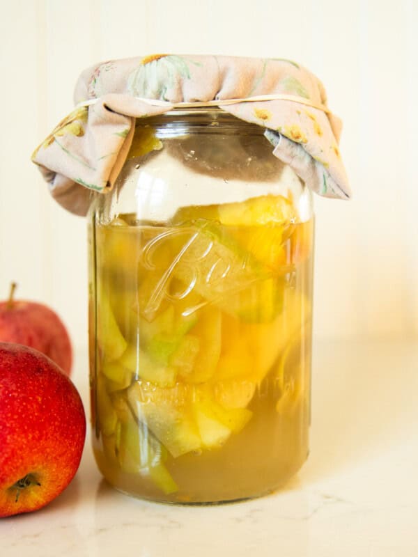 A jar of apple cider vinegar with a cheesecloth on the top and apples next to it.