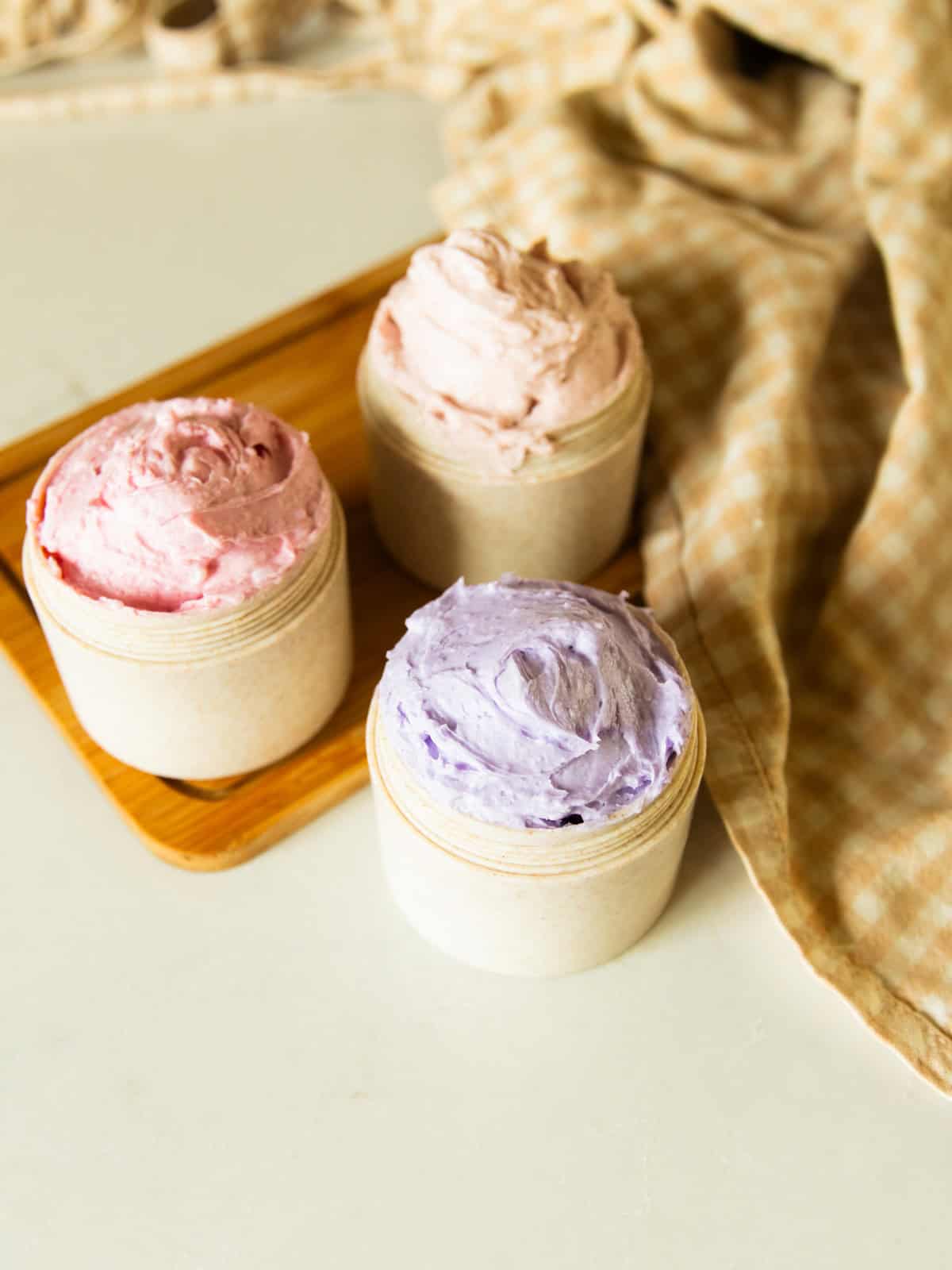 Three jars of foaming sugar soap in three different colors, on a wooden board with a towel. 