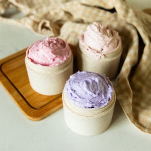 Three jars of different-colored whipped sugar soap on a wooden cutting board with a towel.